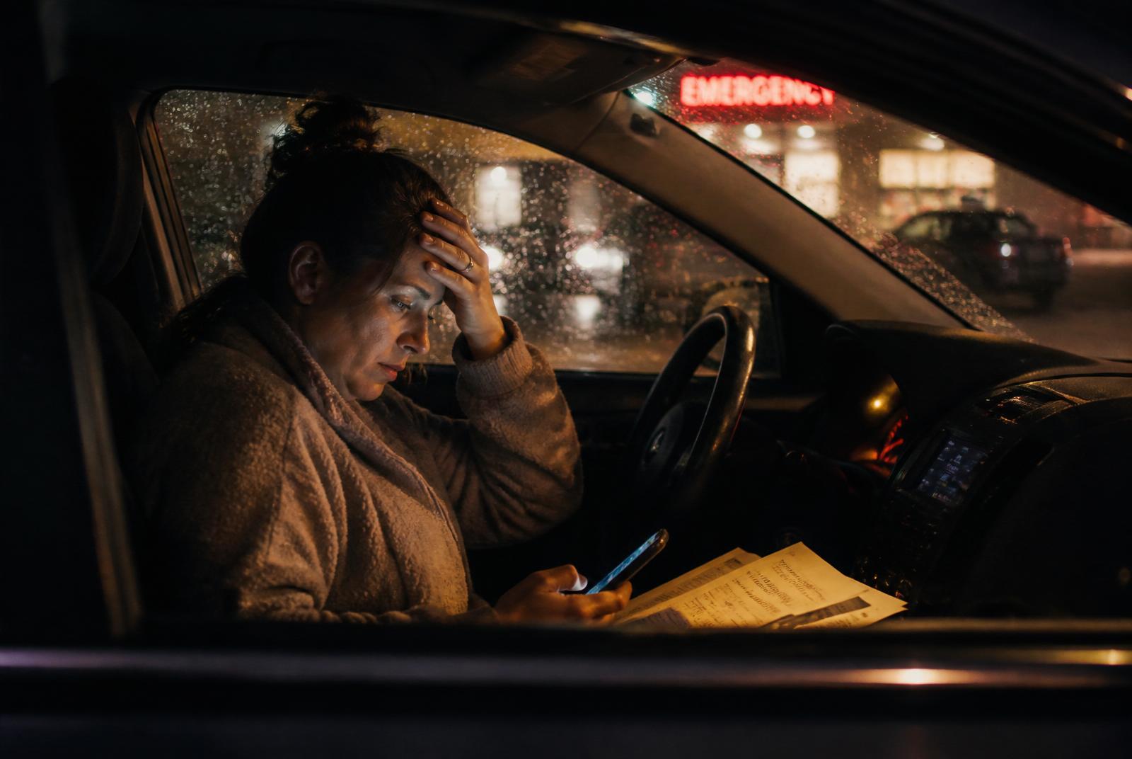 A woman alone in her car in a hospital parking lot at night, hospital paperwork in her lap.
